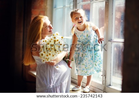 A young pretty pregnant woman with long blonde hair hugging her little cute daughter standing on the windowsill at home. Happy family concept. Parents and kids