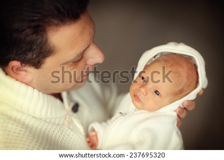 Happy family: a close up portrait of a handsome man in white clothes holding his cute newborn baby in a white suit and looking at him with admiration.