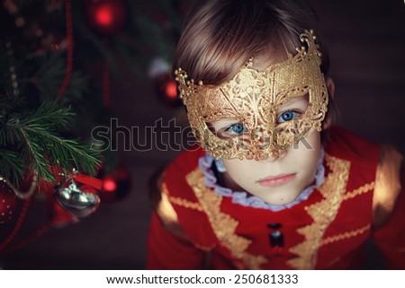A portrait of a little handsome boy in masquerade costume and a carnival mask against the decorated Christmas tree. Fancy-dress ball.