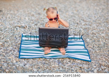 A cute little happy girl in pink sunglasses sitting on the striped blue and white mat with a laptop on the beach on a warm summer day. Holiday on a seashore concept. Funny kids. Sea. Ocean.