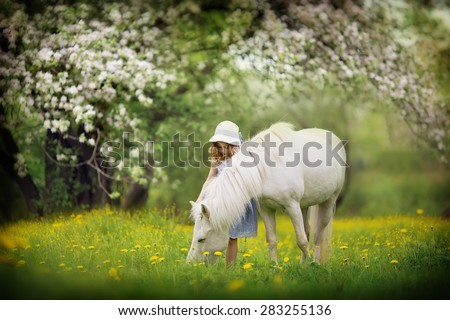 A cute little girl in a white hat caressing her little pony in the blooming garden on a sunny spring day. Kids in the country.