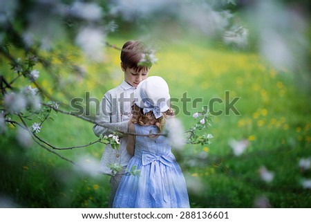 A cute little girl in an elegant white hat and light blue dress and a cute young boy embracing in the blooming garden on a sunny spring day. Kids in the country.