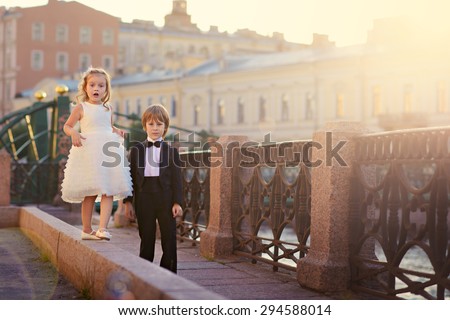 An elegant handsome little boy in a tail-coat and his pretty little girlfriend walking along a beautiful old bridge at sunset on a sunny warm summer evening. Kids in the city. Saint-Petersburg. Russia