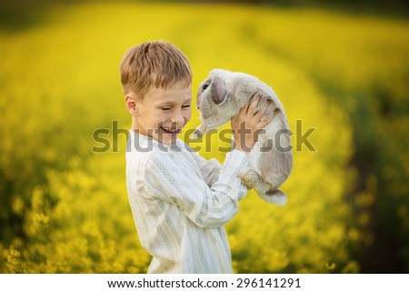 A young cute boy in a white shirt holding a tame rabbit in the field of flowering yellow mustard on a sunny summer day. Kids and nature. Children in country. Beautiful flora