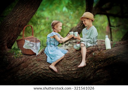 Two cute little kids sitting on a huge brunch of the tree and making up a picnic on a sunny summer day. Children in the country.