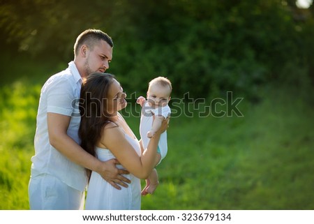 Happy family together: beautiful young parents in white clothes walking with their new-born daughter in the park on a sunny summer day.