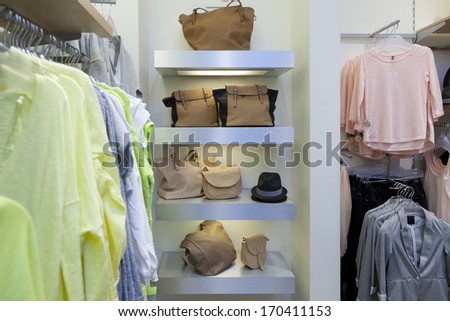 Blouses, shirts hanging on rack in retail shop interior. Bags, handbags, shoulder bags on shelf.
