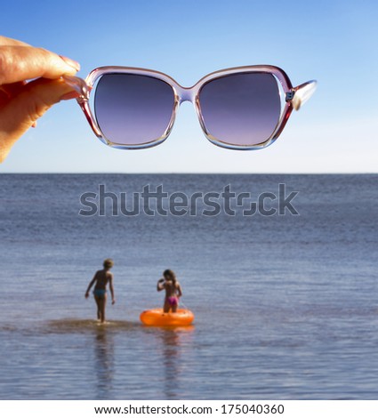 Woman hand holding sunglasses in air over water. Kids in water with inflatable life ring.