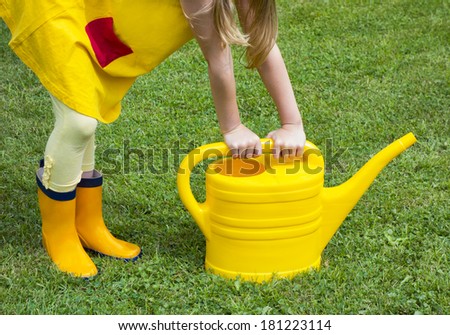 Hands holding watering-can. Closeup. Yellow clothes and rubber boots. Green grass.