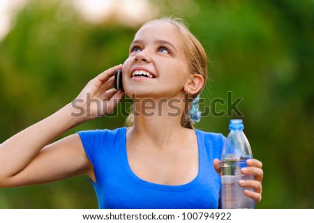 Smiling teenager girl in blue dress holding water bottle and talking on cell phone in green summer park.