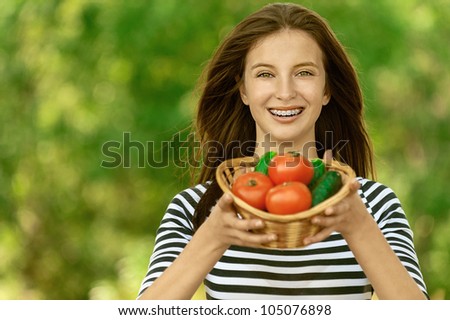 Young attractive woman holding basket of tomatoes and cucumbers, against green of summer park.