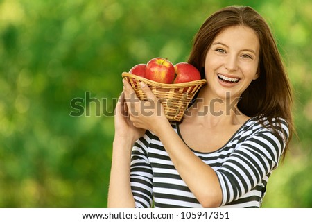 Young attractive woman holding basket with apples, against green of summer park.