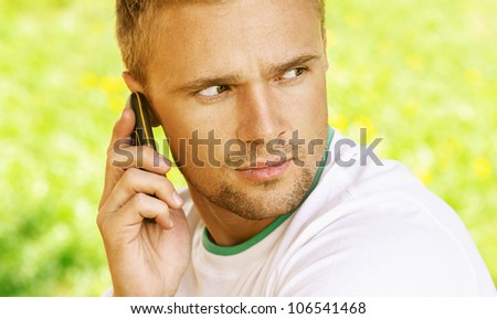 Pretty young man sitting on wooden bench and talk on mobile phone, against green of summer park.