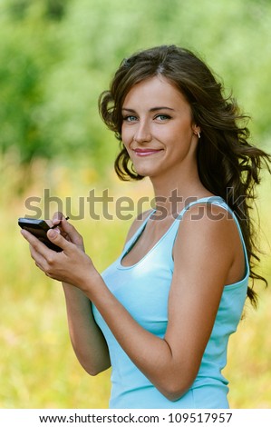 Beautiful young woman close-up in blue shirt is typing with stylus on device, against green of summer park.