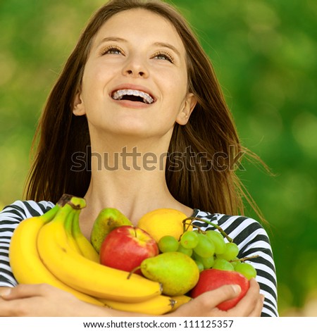Young attractive woman holding apples, pears, grapes and bananas, against green of summer park.