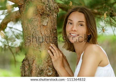 Dark-haired smiling beautiful young woman in white blouse near trunk tree, against green of summer park.