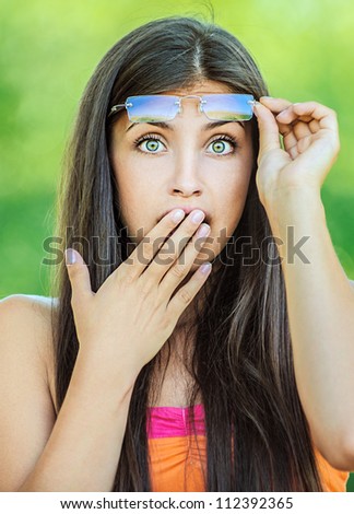 Portrait of young beautiful woman picks up glasses and closes his hand in surprise mouth, on green background summer nature.