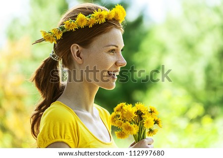Portrait of red-haired smiling beautiful young woman in yellow blouse with wreath and bouquet of dandelions, against green of summer park.