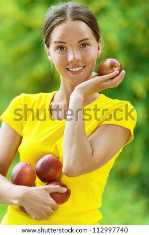 Portrait of dark-haired smiling beautiful young woman in yellow blouse with apples, against green of summer park.