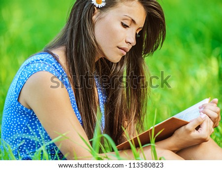 Portrait of young beautiful woman sitting on grass and reading book, on green background summer nature.