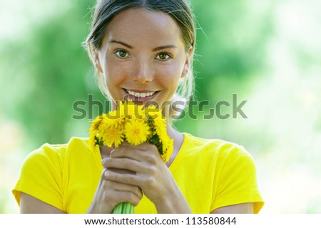 Portrait of dark-haired smiling beautiful young woman in yellow blouse with a bouquet of dandelions, against green of summer park.