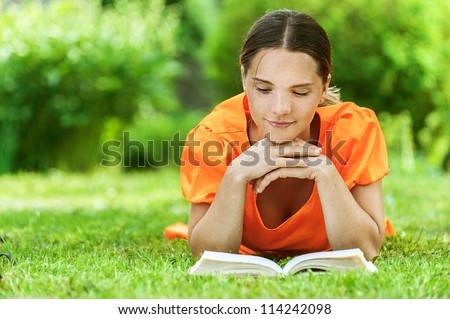 Dark-haired smiling beautiful young woman in orange blouse lying on grass and reading book, against green of summer park.