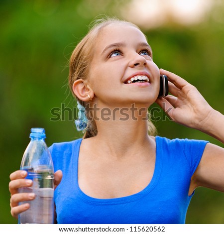 Smiling teenager girl in blue dress holding water bottle and talking on cell phone in green summer park.