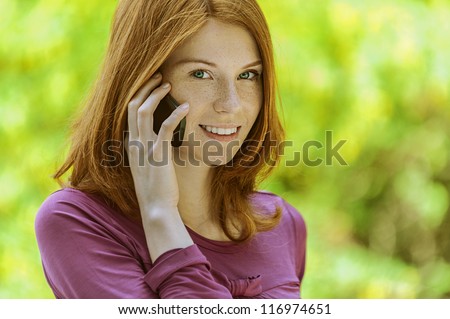 Portrait of red-haired smiling beautiful young woman with mobile phone, against green of summer park.