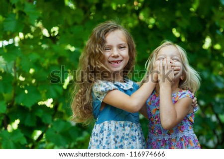 Two sisters playing - with one hand covers mouth of another, against background of summer city park.