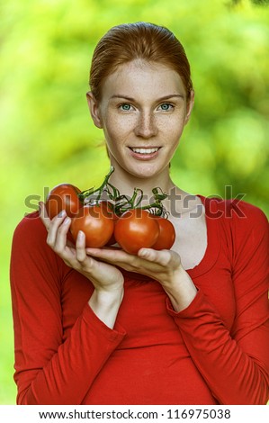 Portrait of red-haired smiling beautiful young woman in red blouse with tomato, against green of summer park.