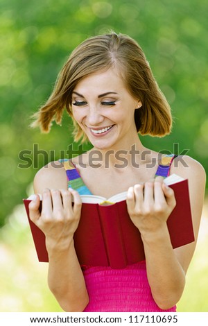 Portrait of smiling beautiful young woman reading red book, against green of summer park.