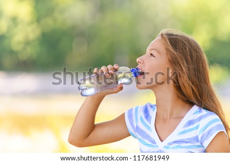 Beautiful teenage girl in blue blouse drinks water from bottle, against green of summer park.