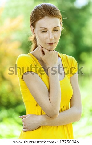 Portrait of red-haired pensive beautiful young woman in yellow blouse, against green of summer park.