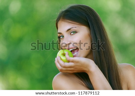 Nice dark-haired funny young woman bites off an apple, against background of summer green park.