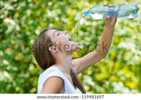 Beautiful young woman drinks water from bottle, against green of summer park.