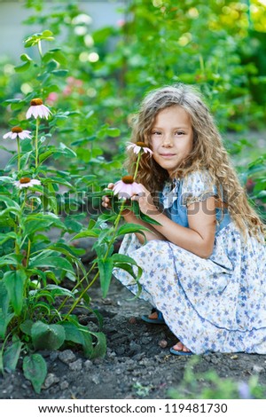 Portrait of beautiful smiling little girl with curly hair about growing flowers, against background of summer garden.