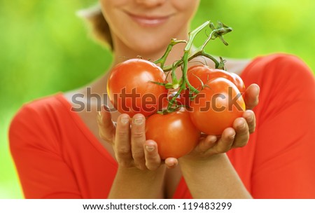 Portrait of dark-haired smiling beautiful young woman in red blouse with tomato, against green of summer park.