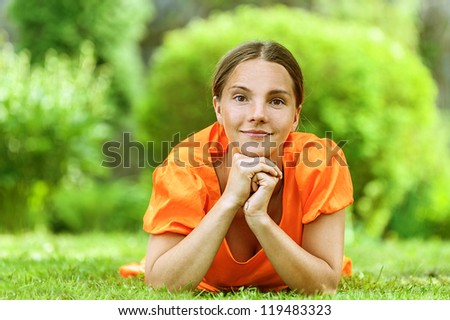 Dark-haired smiling beautiful young woman in orange blouse lying on grass, against green of summer park.