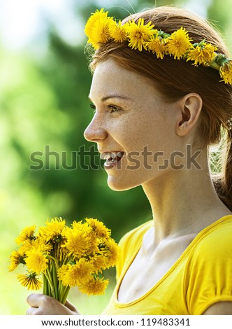 Portrait of red-haired smiling beautiful young woman in yellow blouse with wreath and bouquet of dandelions, against green of summer park.