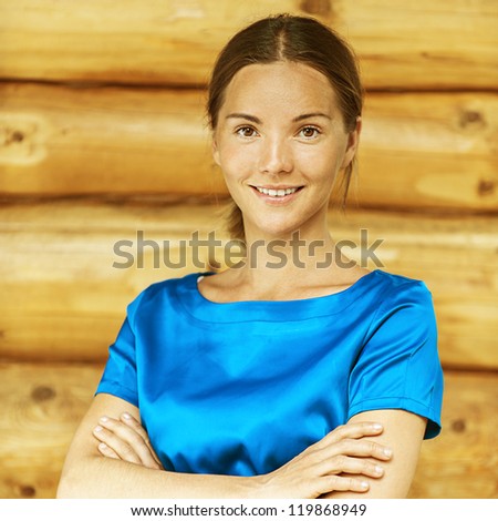 Portrait of dark-haired beautiful young woman in blue blouse, against background of log hut