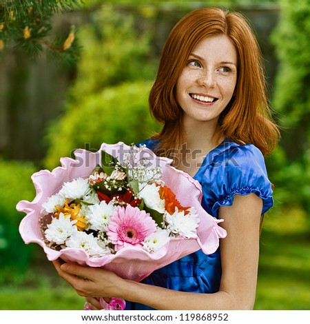 Red-haired smiling beautiful young woman in blue blouse with bouquet of flowers, against green of summer park.