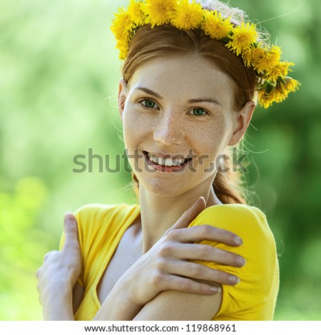 Portrait of red-haired smiling beautiful young woman in yellow blouse with wreath of dandelions, against green of summer park.