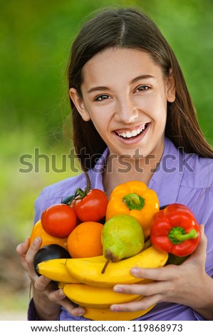 Smiling teenage girl holding banana, peppers, pears and oranges, against green of summer park.