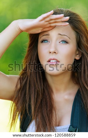 Beautiful young woman with hand closes eyes from sun and looks into distance, against background of summer green park.