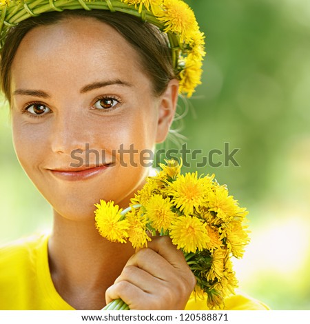 Portrait of dark-haired smiling beautiful young woman in yellow blouse with wreath and bouquet of dandelions, against green of summer park.