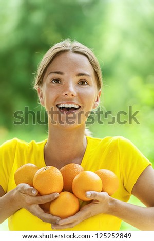 Portrait of dark-haired smiling beautiful young woman in yellow blouse with oranges, against green of summer park.