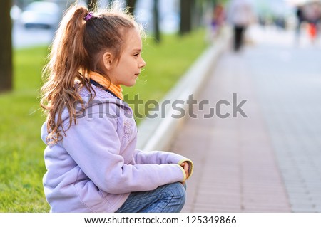 Beautiful little girl in jacket sitting on paving-stone curb profile, against background of city street.