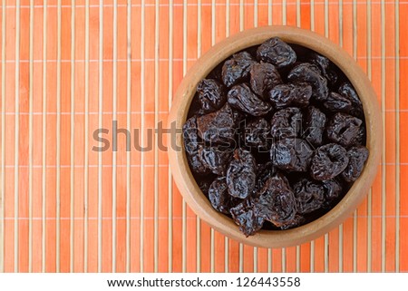 Dried prunes in wooden bowl on orange bamboo table cloth.