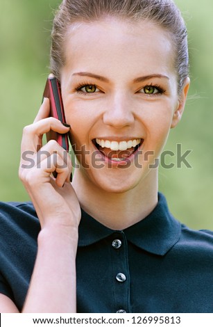 Beautiful smiling young woman in dark blouse talking on mobile phone, against green of summer park.