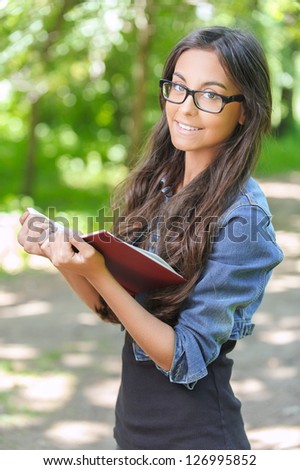 Portrait of beautiful smiling dark-haired young woman with book in denim jacket, against summer green park.
